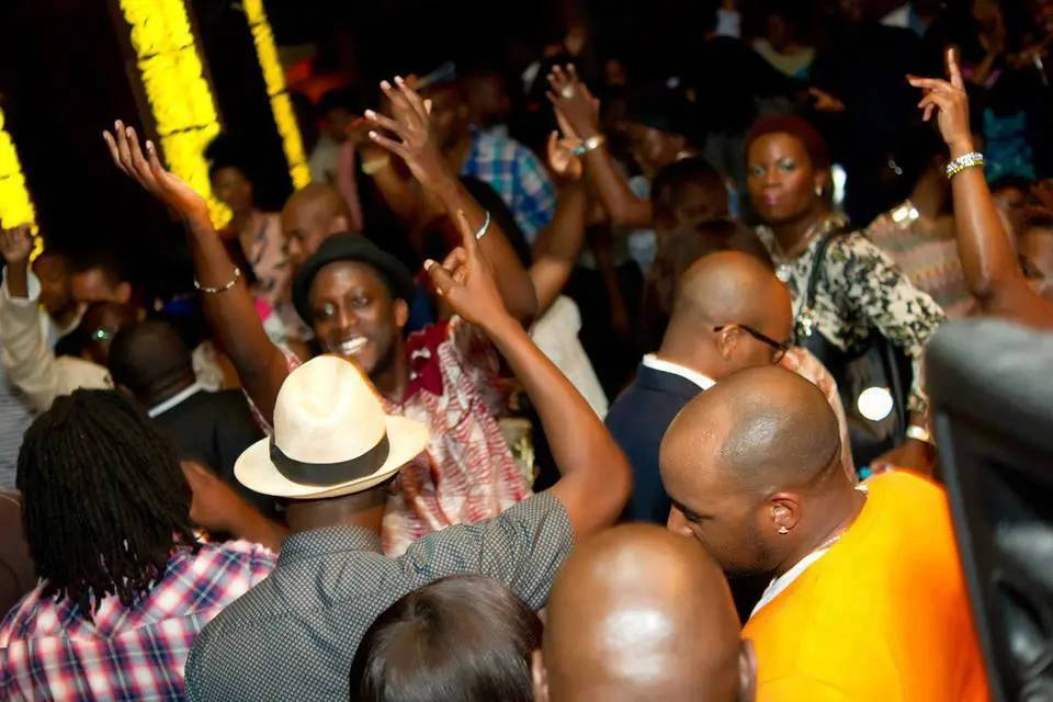 A crowded indoor party with people dancing and DJ Sola keeping the energy high, all under dim lights in casual and semi-formal attire.