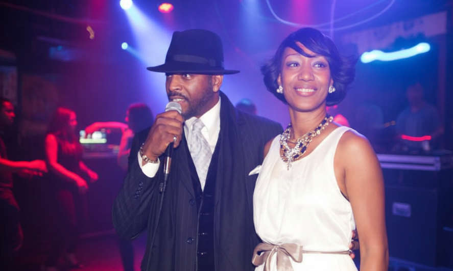 A top NYC MC Euneek of Maestro Beats DJ & Entertainment Service - in a suit and hat speaks into a mic beside a celebrant, a Mastrobeats DJ Service corporate client in a white dress