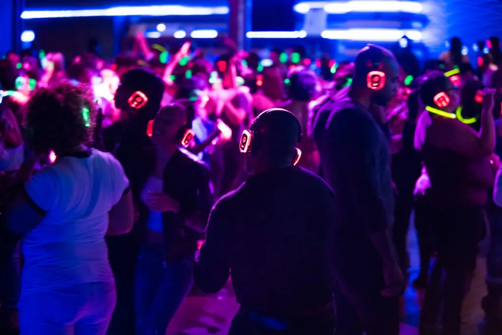 A crowd, including Alex, wears glowing headphones and dances at a silent disco with blue and pink lights in a dimly lit venue.