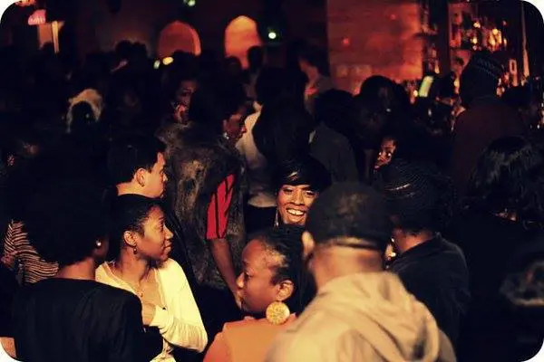 MaestroBeats indoor party with people socializing, some smiling and talking, in dim lighting as DJ Sbu sets the Amapiano vibe.