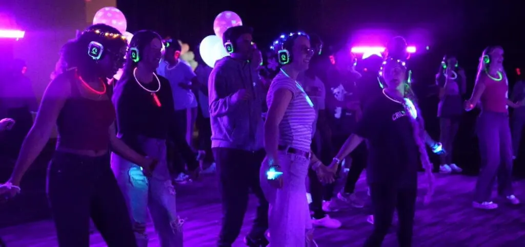 A group of people, including Alex, wear headphones and glow accessories while dancing in a dark room with purple and blue lights.