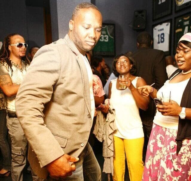 Afropop dances at a social event as people smile and laugh nearby. Framed jerseys hang behind him while an Amapiano DJ plays music.